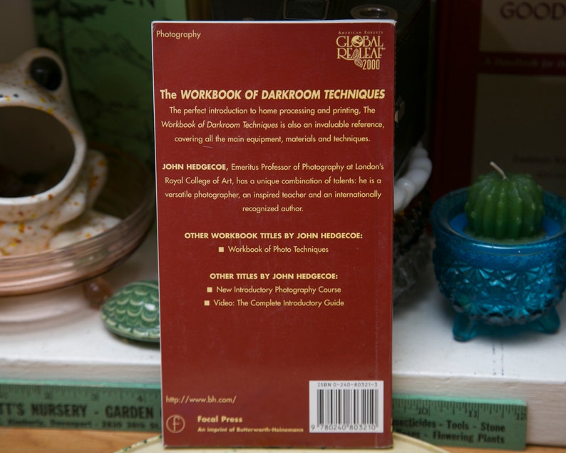 Op de afbeelding: Een rood boek met de titel "The Workbook of Darkroom Techniques" van John Hedgecoe. De tekst beschrijft het als een introductie tot thuisverwerking en afdrukken. Het boek wordt gepresenteerd op een plank met andere decoratieve items.