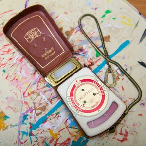 May include: A white and red light meter with a black dial and a gold trim. The meter is open and the top half is a brown leather case with the text "ZEISS IKON STUTTGART GERMANY" on it. The meter is on a white surface with colorful paint splatters.