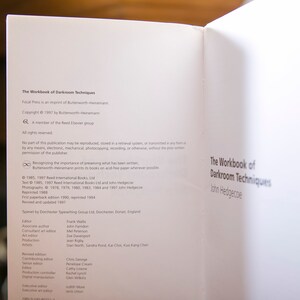 Op de afbeelding: Open boek met de titel "The Workbook of Darkroom Techniques" van John Hedgecoe. De pagina toont copyrightinformatie, uitgeversgegevens en een lijst met bijdragers. Het boek is gedrukt op wit papier.