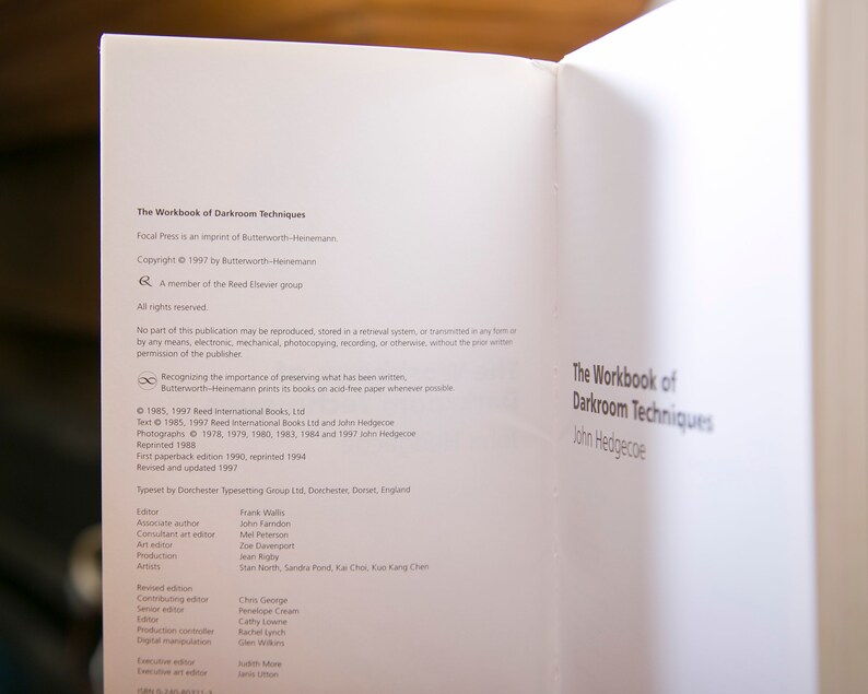 Op de afbeelding: Open boek met de titel "The Workbook of Darkroom Techniques" van John Hedgecoe. De pagina toont copyrightinformatie, uitgeversgegevens en een lijst met bijdragers. Het boek is gedrukt op wit papier.
