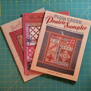 May include: Three quilting pattern books with red, white, and blue covers. The top book is titled "Plum Creek Prairie Sampler" with the subtitle "27 Projects to Quilt for Your Home". The middle book is titled "More Time" and the bottom book is titled "Hearts".