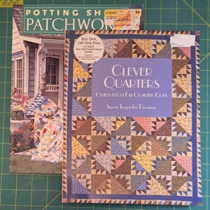 May include: Two books on a green surface. The book on the right is titled "Clever Quarters" with a quilt pattern on the cover. The book on the left is titled "Potting Shed Patchwork". A sticker on the cover says "Buy One, Get One Free".