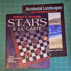 May include: Two quilting books are displayed on a green cutting mat. The book "Stars A La Carte" by Bethany S. Reynolds features a checkerboard design with star patterns. The other book is titled "Accidental Landscapes" by Karen Eckmeier.