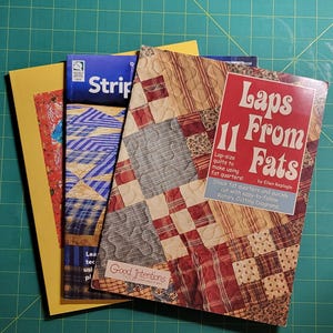 May include: Three quilting books stacked on top of each other. The top book is red and white with the title "Laps 11 From Fats" by Ellen Replagle. The middle book is blue and white with the title "Good Intentions". The bottom book is yellow with the title "Strips".