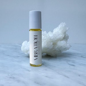 Pode incluir: Um frasco branco com rolo contendo um líquido amarelo. O frasco está etiquetado com "VIBRATION HIGH FREQUENCY ROLLER". O frasco está sobre uma superfície de mármore branco.