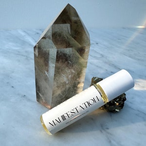 May include: A clear quartz crystal point and a small pyrite cluster are next to a roller bottle. The roller bottle has a white cap and the words "MANIFESTATION" and "REALITY CREATION ROLLER" printed on it.