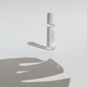 May include: A white roller bottle with a white label that reads "BEE BALM + ASH MOMO POSTPARTUM EMOTIONS".