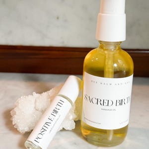 May include: A clear glass pump bottle of Sacred Birth massage oil and a clear glass roller bottle labeled Positive Birth labor support roller, both featuring white labels with black text, positioned in front of a white crystalline mineral cluster.