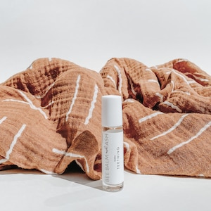 Op de afbeelding: Een witte roll-on fles met een label waarop "Baby Bee Balm + Lash Teething" staat. De fles staat op een bruin-wit gestreepte stof.