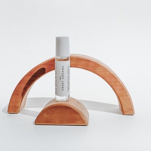 May include: A white roller bottle with a label that reads "child" and "SWEET DREAMS" on a wooden rainbow arch. The bottle is labelled "BELL BALM + ASH" and contains organic coconut oil & essential oils.
