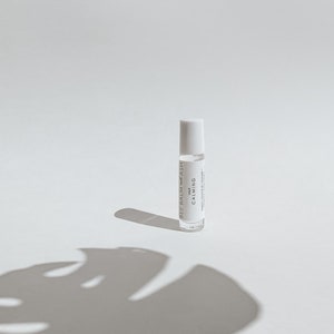 May include: A clear glass roll-on bottle with a white cap, labeled "BEE BALM ASH" and "CALMING." The bottle casts a shadow of a large leaf on a white surface. The product is likely a calming essential oil.