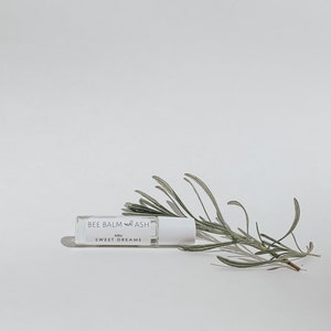 Könnte beinhalten: Eine weiße Roll-on-Flasche mit dem Text "BEE BALM and ASH" und "baby SWEET DREAMS" auf dem Etikett. Die Flasche liegt auf einer weißen Oberfläche mit einem Zweig grünen Rosmarins.