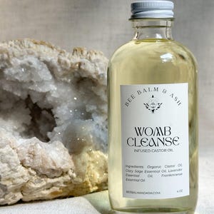 以下が含まれることがあります： 白いキャップが付いた透明なガラス瓶で、「Womb Cleanse Infused Castor Oil」と書かれたラベルが付いています。 ラベルには、オーガニックヒマシ油やラベンダーエッセンシャルオイルなどの成分が記載されています。 ボトルは118 mlです。 その隣には白とベージュのジオードがあります。