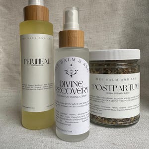 May include: Three Bee Balm and Ash postpartum care products: a massage oil, a perineal spray, and a herbal sitz bath blend. The bottles have wooden caps and white labels with black text. The massage oil is in a clear bottle with a yellow liquid.