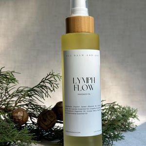 Op de afbeelding: Een massageolie fles van matglas met een houten dop en pomp. Het etiket luidt "LYMPH FLOW MASSAGE OIL". De fles is omgeven door groen en dennenappels. De olie heeft een lichtgele kleur.