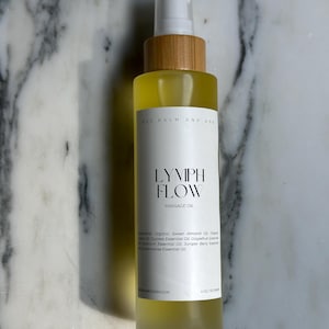 Op de afbeelding: Een heldere glazen fles massageolie met een wit etiket met de tekst "LYMPH FLOW MASSAGE OIL". De fles heeft een houten dop en een witte sproeikop. De olie is geel en de fles bevat 118 ml.