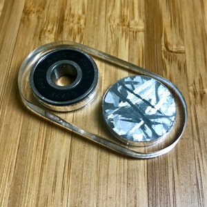 May include: A clear plastic fidget toy with a black and silver bearing and a silver and gray patterned disc.