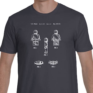 May include: A dark gray t-shirt with a white print of a Lego figure patent drawing. The drawing shows the figure in different positions and includes the text "U.S. Patent: Dec. 23, 1977", "FIG. 1", "FIG. 2", "FIG. 3", "FIG. 4", and "FIG. 5".