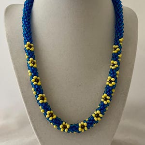 May include: A beaded necklace with a blue base and yellow flower-like accents. The necklace is displayed on a white mannequin bust. The beads are small and tightly woven, creating a textured appearance. The design features a repeating pattern of blue and yellow.