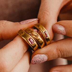 Puede incluir: Tres anillos de oro con piedras preciosas rosas y transparentes. Los anillos están apilados uno encima del otro y están siendo sostenidos por una mano con esmalte de uñas rosa.