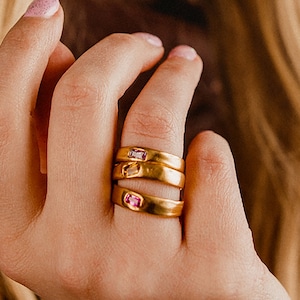 May include: Three gold rings with pink gemstones stacked on a finger. The rings are simple and elegant, with a minimalist design. The gemstones are small and delicate, adding a touch of color to the rings. The rings are perfect for everyday wear or for a special occasion.
