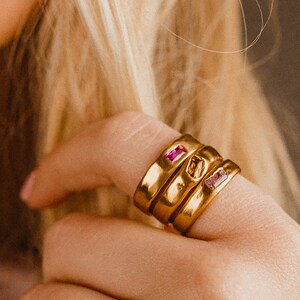 Puede incluir: Tres anillos de oro con piedras preciosas rosas apilados en un dedo. Los anillos son simples y elegantes, con un acabado liso y pulido.