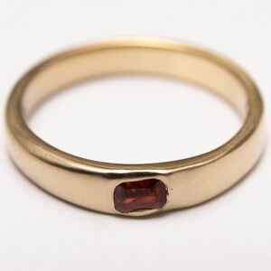 May include: A simple gold band ring with a small, rectangular, red gemstone set in the center.