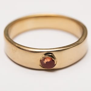 Puede incluir: Un anillo de banda de oro con una sola piedra roja engastada en el centro.
