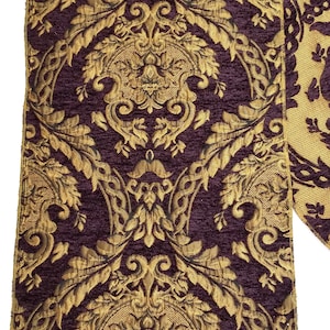 May include: A decorative table runner with a rich, ornate design. The runner features a deep purple background with a gold, damask-style pattern. The fabric has a textured appearance, and the design includes floral and scroll motifs.