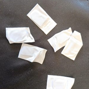 May include: Six white fabric pouches with a small opening on one side. The pouches are folded and arranged on a dark grey surface.