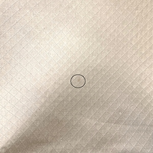 May include: A close-up of a beige fabric with a diamond pattern. The fabric has a small, light brown stain in the center.