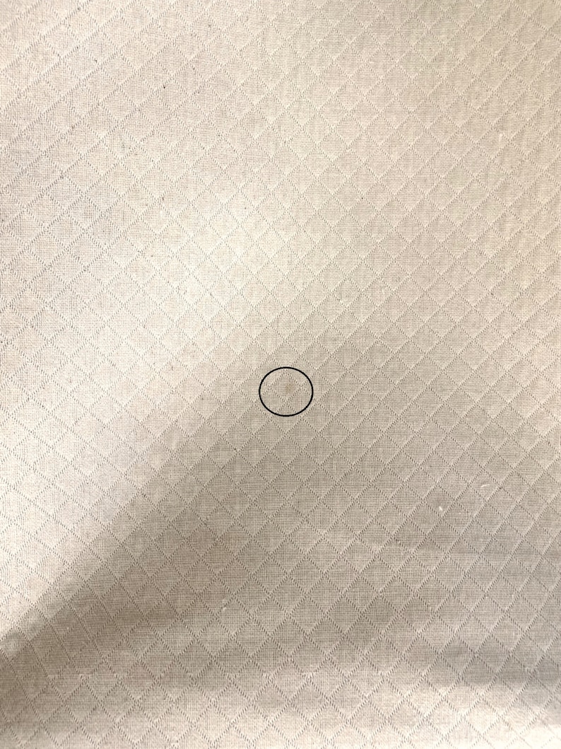 May include: A close-up of a beige fabric with a diamond pattern. The fabric has a small, light brown stain in the center.