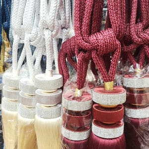 May include: A collection of decorative tassel tiebacks in various colours, including navy blue, white, and burgundy. Each tassel features a layered design with a beaded trim and a long, flowing fringe.