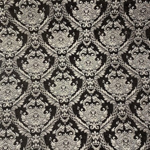 May include: Close-up of a black and silver damask fabric. The pattern features a repeating design of ornate floral and scroll motifs in a metallic silver color against a dark background. The fabric has a textured appearance.