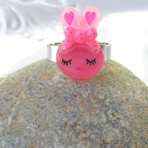 K&ouml;nnte beinhalten: Ein rosa Hasenring mit einem silbernen Band. Der Hase hat eine Schleife, herzf&ouml;rmige Ohren und geschlossene Augen mit Wimpern. Der Ring ist auf einem grauen Stein platziert.