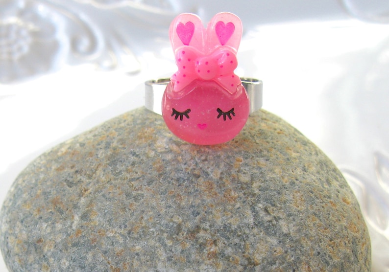 K&ouml;nnte beinhalten: Ein rosa Hasenring mit einem silbernen Band. Der Hase hat eine Schleife, herzf&ouml;rmige Ohren und geschlossene Augen mit Wimpern. Der Ring ist auf einem grauen Stein platziert.