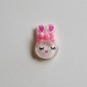 K&ouml;nnte beinhalten: Ein klarer, hasenf&ouml;rmiger Schmuck mit einer rosa Schleife und herzf&ouml;rmigen Ohren. Der Hase hat geschlossene Augen mit Wimpern und ein kleines rosa Herz als Nase. Das Design ist vor einem wei&szlig;en Hintergrund.