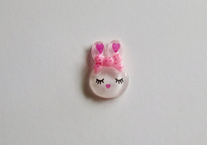 K&ouml;nnte beinhalten: Ein klarer, hasenf&ouml;rmiger Schmuck mit einer rosa Schleife und herzf&ouml;rmigen Ohren. Der Hase hat geschlossene Augen mit Wimpern und ein kleines rosa Herz als Nase. Das Design ist vor einem wei&szlig;en Hintergrund.