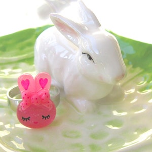 K&ouml;nnte beinhalten: Eine wei&szlig;e Keramik-Hasenfigur und ein rosa Hasen-Ringhalter mit einem silbernen Ring, auf einer gr&uuml;n-wei&szlig;en Keramikschale. Der Ringhalter hat eine Schleife und herzf&ouml;rmige Ohren.