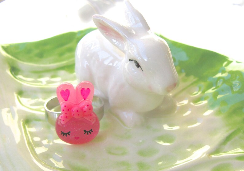 K&ouml;nnte beinhalten: Eine wei&szlig;e Keramik-Hasenfigur und ein rosa Hasen-Ringhalter mit einem silbernen Ring, auf einer gr&uuml;n-wei&szlig;en Keramikschale. Der Ringhalter hat eine Schleife und herzf&ouml;rmige Ohren.