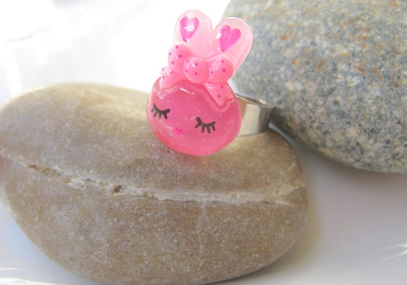 K&ouml;nnte beinhalten: Ein rosa Hasenring mit einem silbernen Band. Der Hase hat eine Schleife und herzf&ouml;rmige Ohren. Der Ring liegt auf einem hellbraunen Stein, mit einem grauen Stein im Hintergrund.