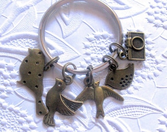 May include: A silver-colored metal keychain with several bronze-colored bird charms and a small camera charm. The bird charms include a variety of designs, such as a bird with a dotted pattern and birds with outstretched wings. The keychain is on a white textured surface.