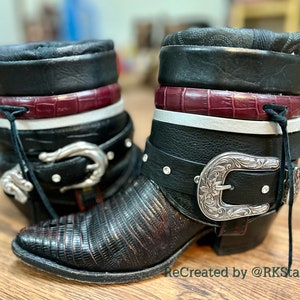 May include: A pair of black leather ankle boots with silver buckles and a burgundy crocodile-embossed leather trim. The boots have a white stripe and a black leather strap with a silver buckle. The boots are on a wooden surface. ReCreated by @RKStarJewelry