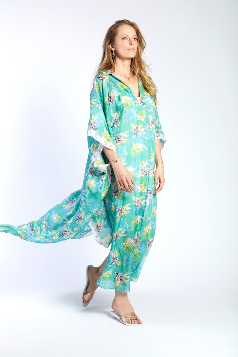 May include: A woman wearing a long, flowing green and white floral print kaftan with a white trim. She is wearing white sandals and has long blonde hair.