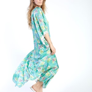 May include: A woman wearing a long, flowing, teal blue and green floral print dress with white lace trim on the sleeves. She is wearing gold sandals.