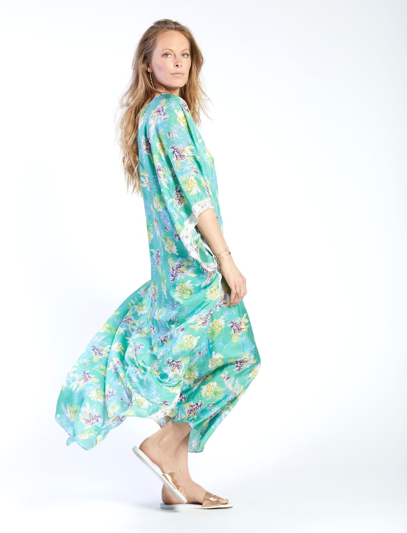 May include: A woman wearing a long, flowing, teal blue and green floral print dress with white lace trim on the sleeves. She is wearing gold sandals.