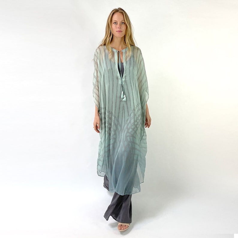 May include: A long, flowing, sheer dress in shades of seafoam green and gray. The dress features vertical stripes and a subtle, patterned design. It has a keyhole neckline with a tassel detail and is paired with gray pants.