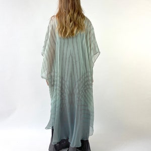 May include: A flowing, light blue and gray patterned kimono-style garment. The sheer fabric features vertical stripes and a subtle gradient effect. The garment is worn over darker, solid-colored pants.