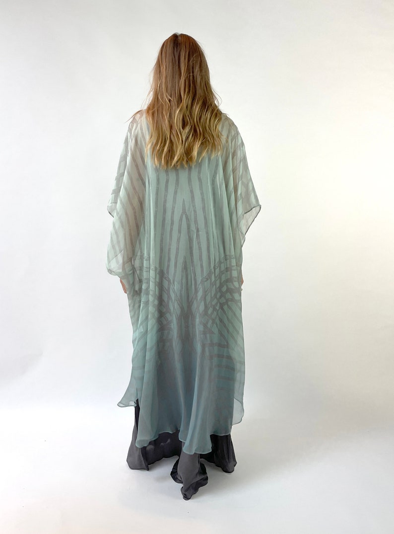 May include: A flowing, light blue and gray patterned kimono-style garment. The sheer fabric features vertical stripes and a subtle gradient effect. The garment is worn over darker, solid-colored pants.