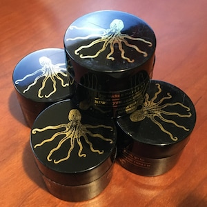 May include: Four black jars with gold octopus designs stacked on a wooden surface. The jars have a shiny, glossy finish.
